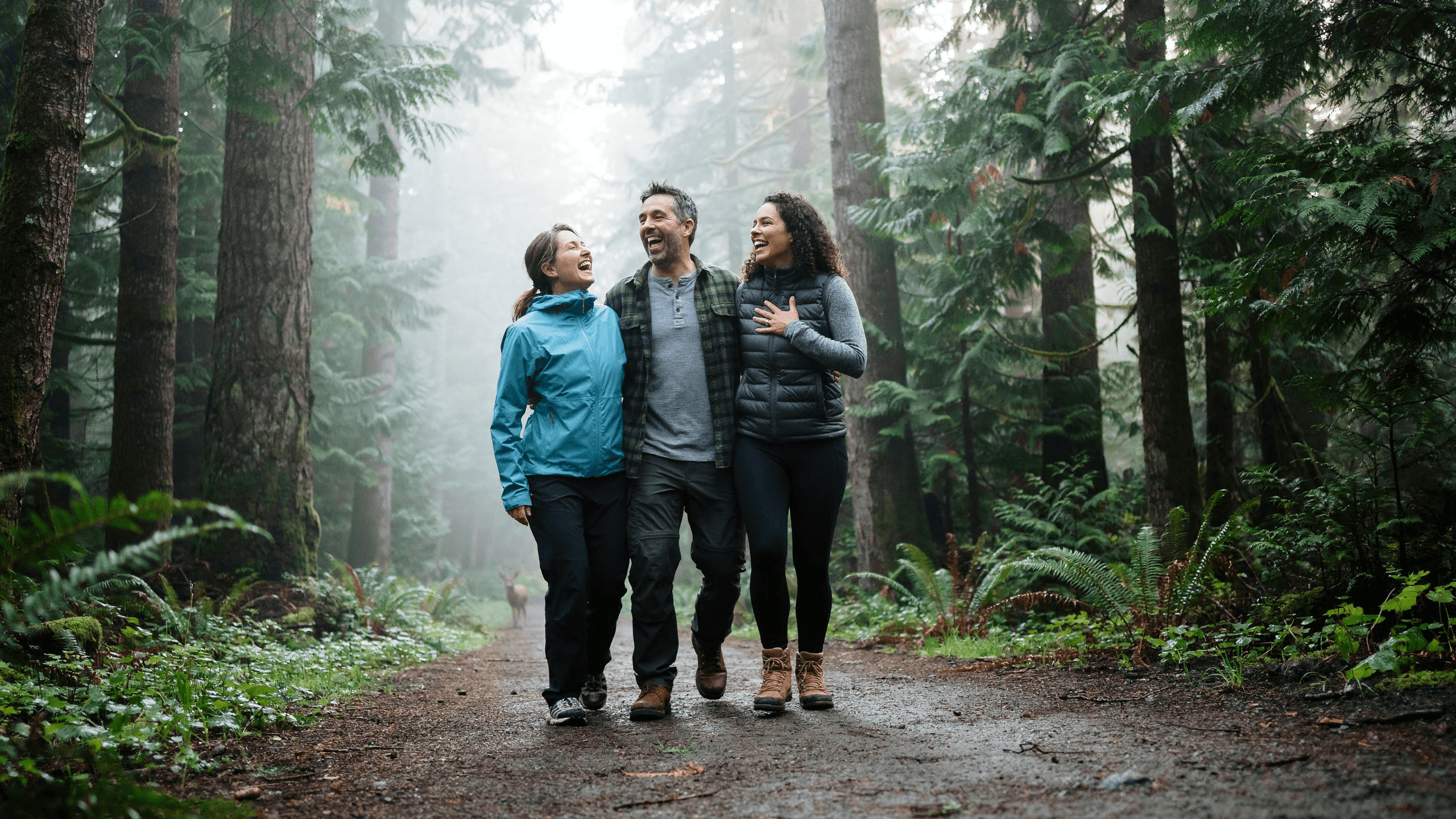 Three friends laughing together on a misty British Columbia forest trail, one wearing a sky blue rain jacket