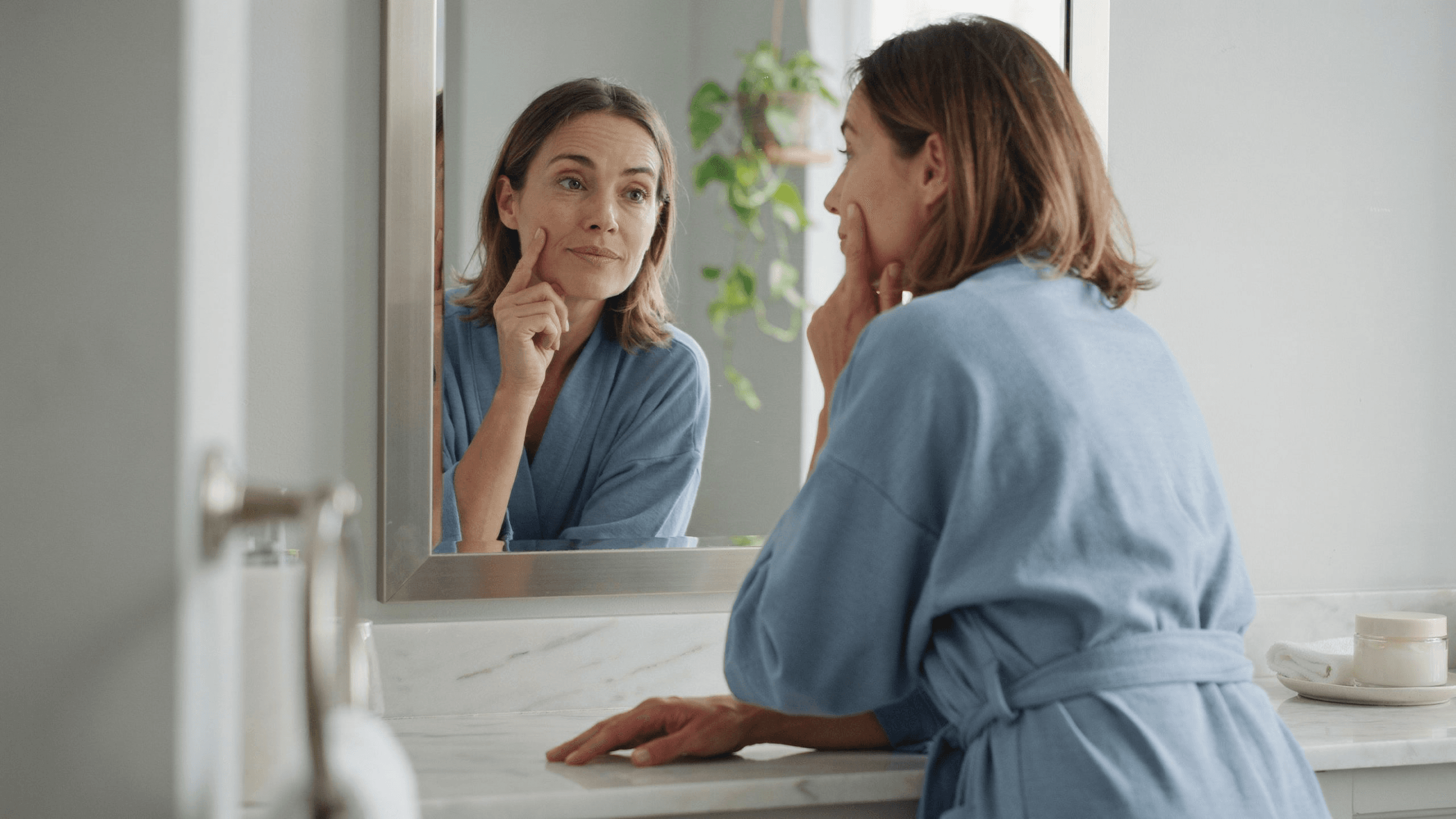 A woman in a sky blue robe leaning toward a bathroom mirror in soft morning light, studying her cheek
