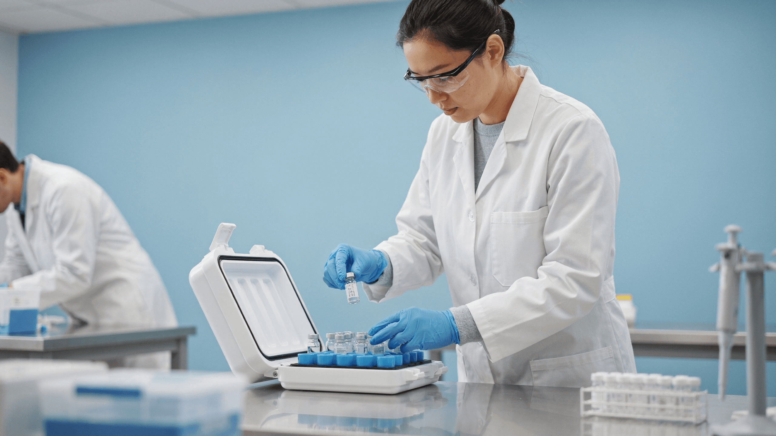 A researcher in safety glasses and blue gloves lifting a small vial out of an insulated case lined with blue cold packs